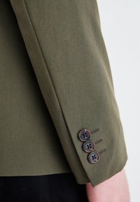 Olive'owy zielony rękaw marynarki z trzema ciemnymi guzikami i detalami przeszyciami. Gładka tkanina, dopasowany krój i wyraźne krawędzie.