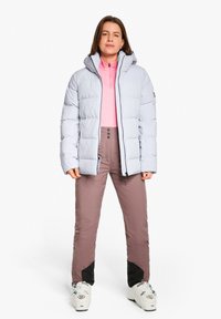 Giacca imbottita grigia chiaro con cappuccio, chiusura lampo frontale e texture trapuntata, abbinata a uno strato base rosa e pantaloni da sci marroni con dettagli neri.