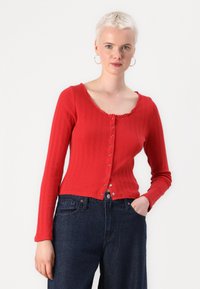 Cardigan rouge à côtes longues avec un col festonné, fermeture à boutons sur le devant et une coupe courte, associé à un jean taille haute bleu foncé.