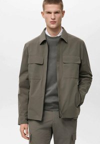 Olivgrüne Jacke mit zwei Brusttaschen, Reißverschluss und einem Kragen, kombiniert mit einem grauen Sweatshirt darunter.