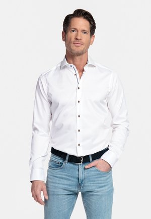 Mann mit kurzen braunen Haaren und leichtem Bart, der ein weißes Hemd mit Knöpfen trägt, das in hellblaue Jeans mit schwarzem Gürtel gesteckt ist, eine Hand in der Tasche.