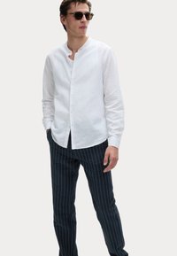 Chemise en lin blanche à col rond, avec un devant boutonné et de longues manches. Associée à un pantalon rayé bleu marine, créant un contraste de textures.