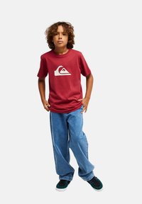 Κόκκινο βαμβακερό T-shirt με λευκό λογότυπο κυματισμού, συνδυασμένο με φαρδιά μπλε τζιν και μαύρα και τιρκουάζ αθλητικά παπούτσια. Χαλαρή εφαρμογή και απλός σχεδιασμός.