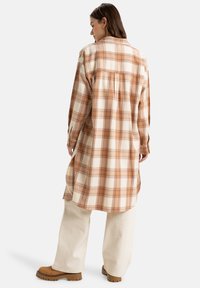 Plaid shirt in beige and orange tones, long sleeves, back pleat detail, curved hem, worn with cream-colored trousers and brown shoes.