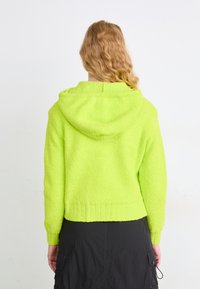 Neon zelena flis dukserica sa teksturiranom tkaninom, kapuljačom i rebrastim rubom. Stilizirano s crnom suknjom, vidljiv je stražnji dio.