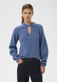 Ženska z svetlo rjavimi lasmi v čopu, oblečena v modro pleteno pulover s ovratnikom in odprtjem, temnimi hlačami, stoji z eno roko v žepu.