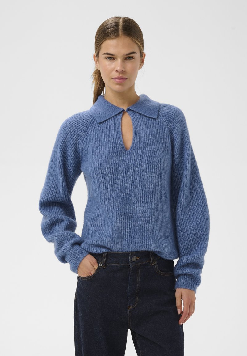 Ženska z svetlo rjavimi lasmi v čopu, oblečena v modro pleteno pulover s ovratnikom in odprtjem, temnimi hlačami, stoji z eno roko v žepu.