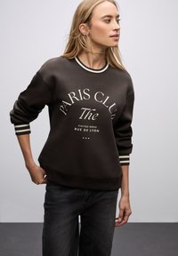 Czarna bluza z grafiką "PARIS CLUB" w kolorze écru. Posiada prążkowany dekolt i mankiety, luźny krój oraz miękką teksturę.