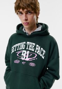 Felpa con cappuccio verde in tessuto morbido con grafica frontale bianca e rosa, con la scritta "SETTING THE PACE" e il numero "91".