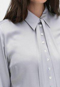 Hellgrau, kragenlose Bluse aus glattem Stoff. Verfügt über eine durchgehende Knopfleiste mit sechs runden Knöpfen und einem eleganten Design.