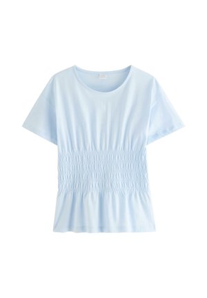 Młoda kobieta w okularach, ubrana w jasnoniebieską marszczoną bluzkę, białe szorty, białe sandały i gumkę do włosów na nadgarstku, stoi rozluźniona.