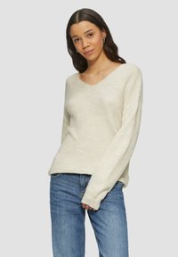 Crème-kleurige gebreide trui met V-hals, lange mouwen, ribbeltextuur en een relaxte pasvorm, gecombineerd met blauwe jeans.