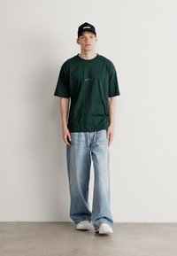 Mørk grønn bomulls t-skjorte med logo foran og løse, lyseblå denimjeans. En svart caps med logo og hvite sneakers fullfører antrekket.
