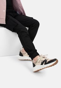Zapatillas con parte superior de malla beige, acentos de cuero negro y una suela gruesa blanca. Pantalones jogger negros con bolsillos laterales.