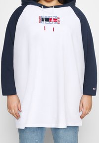 Felpa con cappuccio, corpo bianco e maniche raglan blu navy. Presenta un logo Tommy Jeans ricamato in rosso e blu. Tessuto misto cotone.