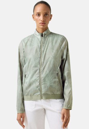 Kobieta w jasnooliwkowej kurtce zapinanej na zamek z subtelnym wzorem, stojąca na tle jednolitej białej ściany, skierowana twarzą do przodu.