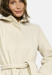Cappotto con cappuccio di colore crema chiaro in tessuto morbido, dotato di cintura, bottoni e dettagli cuciti. Il design include una vestibilità comoda e una superficie strutturata.