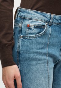 Κοντινό πλάνο ενός ατόμου που φοράει ανοιχτό μπλε τζιν τζιν και καφέ πουλόβερ μακρυμάνικο, με το χέρι του να ακουμπάει δίπλα στο τζιν.