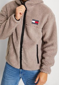 Giacca in pile beige con cappuccio, caratterizzata da una chiusura lampo frontale, tasche laterali e una piccola etichetta con logo sulla parte sinistra del petto.