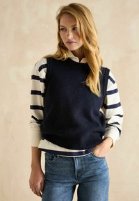 Marineblaues Strickwesten über einem cremefarbenen und marineblauen gestreiften Oberteil, kombiniert mit Jeans. Die Weste hat einen runden Halsausschnitt und einen gerippten Saum.