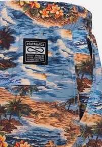 Shorts da bagno con stampa tropicale blu, caratterizzati da palme e onde, con un'etichetta nera. Il materiale è tessuto leggero con una consistenza liscia.