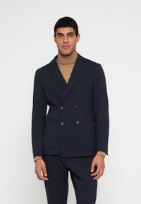 Μπλε ναυτικού τύπου σακάκι διπλής σειράς με δύο μπροστινές τσέπες, κατασκευασμένο από υφασμάτινη υφή, συνδυασμένο με καφέ πουλόβερ με ψηλό γιακά από κάτω.