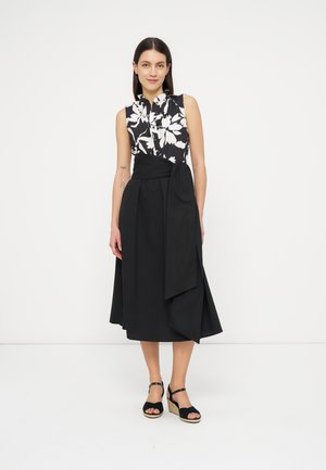 Naine seisab, kandes varrukateta musta-valget lillelist tippu, musta midi-seelikut suure sõlmitud vööga taljes ning musti kiilkontsas sandaale.