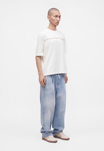 Άνδρας που φοράει λευκό oversized t-shirt, φαρδιά ανοιχτό μπλε τζιν και μπεζ αθλητικά παπούτσια, σε απλό λευκό φόντο.