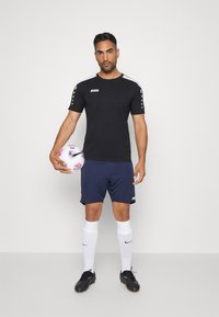 Zwart sportshirt met witte accenten, marineblauwe short, witte sokken en zwarte voetbalschoenen, met een meerkleurige voetbal in de hand.