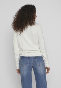 Suéter blanco de punto con mangas largas, dobladillo y puños acanalados, combinado con jeans de mezclilla azul de tiro alto y pierna ancha con bolsillos traseros.