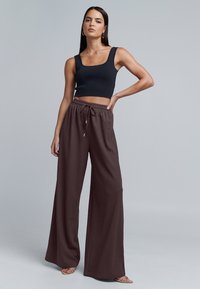 Černý přiléhavý crop top, hnědé široké kalhoty s texturovanou tkaninou a stahovacími šňůrkami v pase, model stojící s jednou rukou na bocích na šedém pozadí.