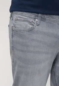 Ανοιχτό γκρι τζιν με πέντε τσέπες, φερμουάρ και ελαφρύ ξεθώριασμα. Το ύφασμα έχει απαλή υφή με λεπτομέρειες ραφών.