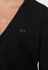 Maglione nero con design a scollo a V, realizzato in tessuto morbido. Presenta un piccolo logo ricamato in grigio sulla parte sinistra del petto.