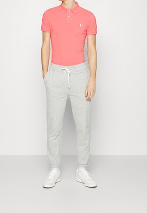 Rode poloshirt met korte mouwen en een kraag, gecombineerd met lichtgrijze joggingbroek met een elastische tailleband en trekkoord. Witte sneakers zichtbaar.