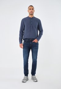 Bărbat în picioare, îmbrăcat cu o cămașă lungă de culoare albastru închis, blugi și adidași gri-silver, având o mână în buzunar.