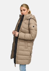 Woman wearing a beige quilted hooded winter coat, partially zipped, with light blue jeans against a plain white background.
