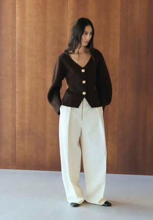 Donna con lunghi capelli scuri che indossa un cardigan marrone a scollo a V con bottoni dorati e pantaloni bianchi larghi, in piedi contro una parete di pannelli di legno.