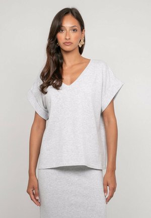 Femme aux longs cheveux foncés portant un haut gris clair à manches courtes en V et une jupe assortie, debout devant un fond uni.