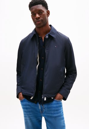 Mand iført en marineblå let jakke over en mørk skjorte med knapper og blå jeans, stående med hænderne i lommerne mod en hvid baggrund.