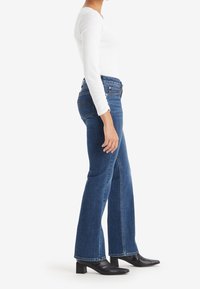 Femme portant un jean évasé bleu taille haute, un haut blanc ajusté à manches longues, et des bottines noires à talons, debout de profil.