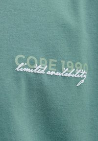 Tissu vert avec texte imprimé "CORE 1990" et texte blanc brodé "disponibilité limitée" qui se chevauche.