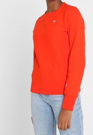 Osoba nosząca jaskrawo pomarańczową, długą bluzę z małym logo na piersi i rękawie, w połączeniu z jasnoniebieskimi dżinsami.