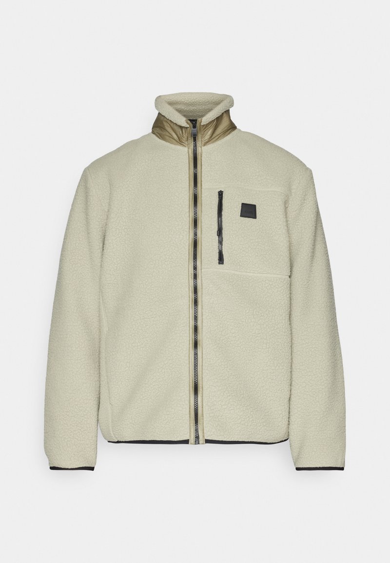 Hellbeige Fleecejacke mit hohem Kragen, Frontreißverschluss und einer Brusttasche. Mit schwarzen Reißverschluss- und Besatzdetails. Weicher Stoff.