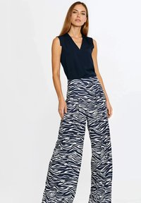 Kobieta w czarnym topie bez rękawów i szerokich spodniach z wysokim stanem w granatowo-białe paski zebry, stoi na jednolitym tle.