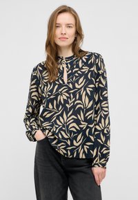 Blouse bleu marine avec un motif de feuilles crème, décolleté en forme de trou de serrure et manches longues. Fabriquée dans un tissu léger et lisse. Associée à un jean foncé.