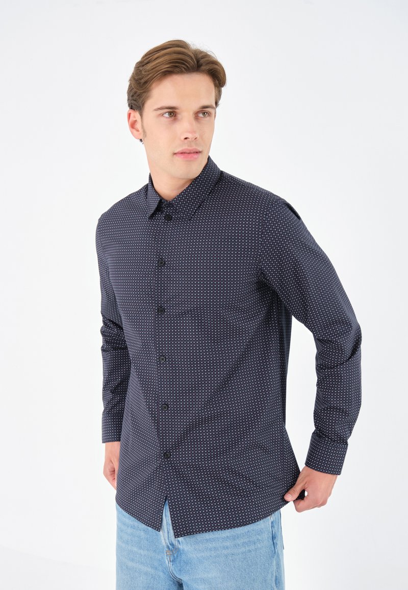 Marineblaues Hemd mit Knöpfen und kleinen weißen Punkten, mittelschwerer Stoff, klassischer Kragen und lange Ärmel, getragen über hellblauen Jeans.