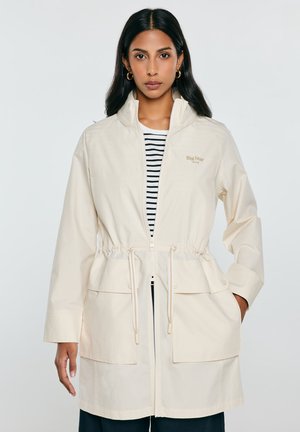 Kobieta ubrana w beżową kurtkę na zamek z troczkami i dużymi kieszeniami na czarno-białą koszulkę w paski, stojąca na jednolitym tle.