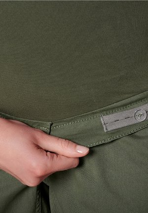 Pantalon vert olive avec une texture lisse et une ceinture visible. Une main serre la ceinture, qui comporte une étiquette gris clair.