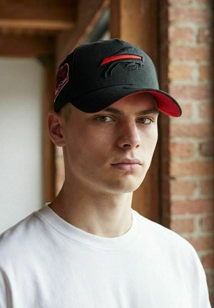 Junger Mann mit schwarzer und roter Baseballkappe mit einem Büffel-Logo und weißem Rundhalsausschnitt, der drinnen nahe einem Fenster mit Ziegelsteinmauer steht.