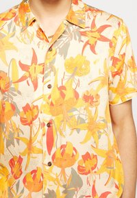 Camicetta a maniche corte con bottoni in un tessuto beige chiaro, caratterizzata da un vivace motivo floreale in tonalità di arancione e giallo con accenti verdi.